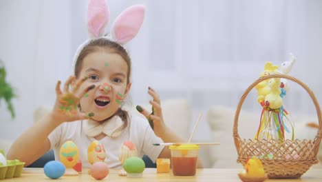 Little-cute-and-adorable-girl-is-smiling-sincerely.-She-takes-an-Easter-egg-and-shows-the-result-of-her-work,-using-her-dirty-of-paints-hands.-Girl-is-going-to-scare-you,-in-a-kind-manner.