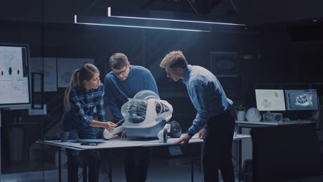 Tarde-en-la-noche-en-el-centro-de-ingeniería-de-robótica-tres-ingenieros-técnicos-hablan-y-trabajan-en-un-prototipo-de-robot-con-ruedas.-En-el-fondo-centro-de-investigación-de-alta-tecnología-con-pantallas-que-muestran-diseño-industrial