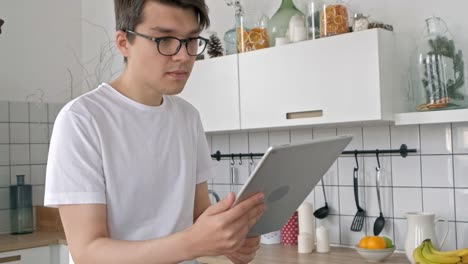 Attractive-man-at-home-using-tablet-in-kitchen-sending-message-on-social-media-smiling-enjoying-modern-lifestyle-wearing-white-shirt