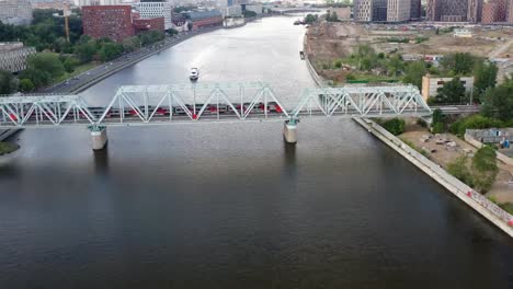 Luftaufnahme-der-Eisenbahnbrücke,-mit-einem-fahrenden-Zug-auf-ihr,-über-den-Fluss,-der-durch-eine-Großstadt-fließt