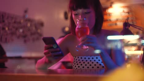 Young-asian-millennial-woman-using-smartphone-and-drinking-beer-in-nightclub-bar,-lifestyle-concept