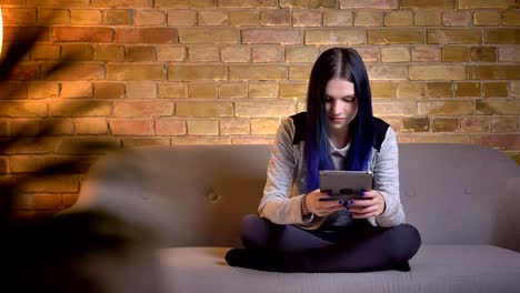 Closeup-shoot-of-young-pretty-caucasian-female-with-dyed-hair-using-the-tablet-and-laughing-cheerfully-while-sitting-on-the-sofa