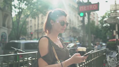 Joven-atractiva-mujer-sonriente-escribiendo-un-mensaje-de-texto-en-su-teléfono-inteligente-en-la-salida-del-metro-en-la-calle,-sosteniendo-su-café,-durante-el-verano-soleado-en-París.-Pecas,-gafas-de-sol,-piercings,-pelo-rojo,-4K.