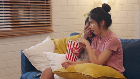 Lesbian-lgbt-women-couple-watching-movie-at-home,-Asian-female-lover-eating-popcorn-scary-and-shock-while-looking-horror-film-together-on-sofa-in-living-room-in-night-concept.