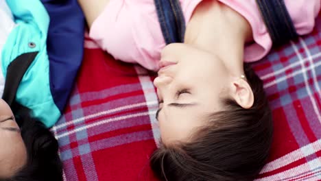 Top-view-of-two-beautiful-diverse-female-students,-Asian-and-Caucasian,-lying-on-plaid-blanket,-looking-at-each-other-and-talking,-closeup-tracking-shot