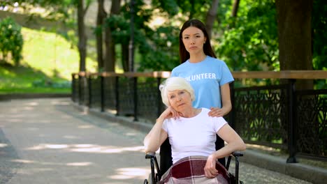 Upset-old-lady-in-wheelchair-and-sad-volunteer-looking-camera-outdoors,-hospice