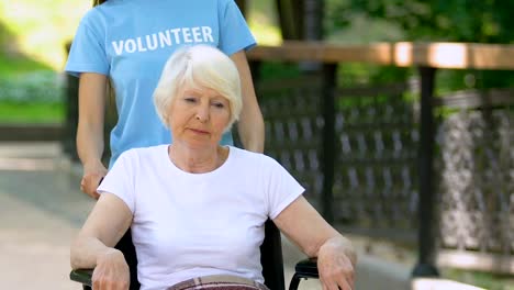 Volunteer-helping-elderly-female-patient-in-wheelchair,-walking-in-hospital-park