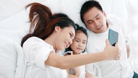 Asiatische-Familie-lacht-und-schaut-in-Smartphone,-während-auf-dem-Bett-im-Schlafzimmer-liegen