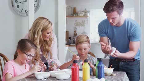 Familie-sitzt-um-Küchentisch-mit-Eltern,-die-Kindern-helfen,-Eier-für-Ostern-zu-malen---in-Zeitlupe-gedreht