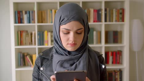 Retrato-de-primer-plano-de-joven-atractiva-estudiante-musulmana-en-hiyab-usando-la-tableta-mirando-la-cámara-en-la-biblioteca-de-la-universidad-en-el-interior