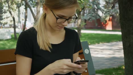 Hübsche-kaukasische-Frau-in-Brille-n.R.-mit-schwarzem-Handy-und-sitzen-auf-der-Bank-im-Stadtpark