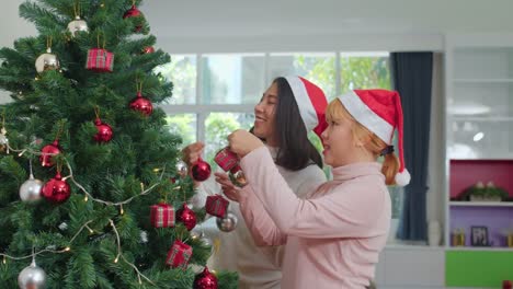 Asian-women-friends-decorate-Christmas-tree-at-Christmas-festival.-Female-teen-happy-smiling-celebrate-xmas-winter-holidays-together-in-living-room-at-home.