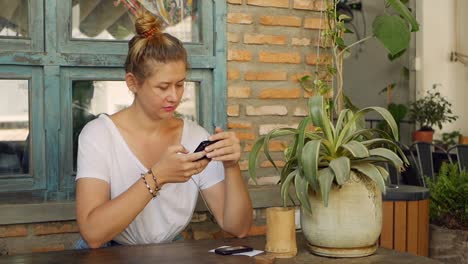 Chica-pensativa-utiliza-un-teléfono-inteligente,-escribe-un-mensaje,-chatea.-Mujer-buscando-información-en-el-teléfono-y-triste.-Sentado-en-el-restaurante-solo.-Hora-del-desayuno.-Concepto-de-tecnología-y-entretenimiento