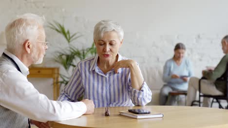 Seguimiento-de-la-toma-media-de-la-pareja-de-ancianos-inteligentes-sentados-en-la-mesa-y-teniendo-discusión,-mujer-mayor-y-anciano-discapacitado-en-silla-de-ruedas-jugando-a-las-cartas-en-el-fondo-en-la-sala-común-de-residencia-de-ancianos