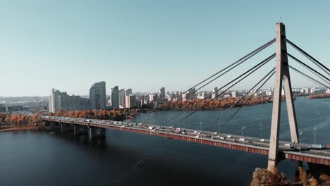 Puente-de-hormigón-cargado-a-través-del-río-en-el-centro-de-la-ciudad.-Tráfico-de-coches-ocupados-en-el-puente,-vista-de-dron-aéreo.---Puente-que-conecta-dos-orillas-de-metrópolis.-Kiev,-Ucrania