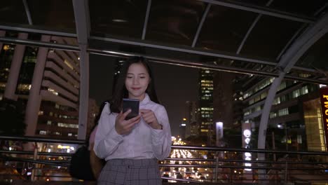 Joven-mujer-asiática-sonriente-de-negocios-usando-teléfono-inteligente-en-el-centro-de-la-ciudad-moderna-en-la-noche-de-Bangkok-Tailandia.-Comunicación-Concept-Technology-por-teléfono-móvil