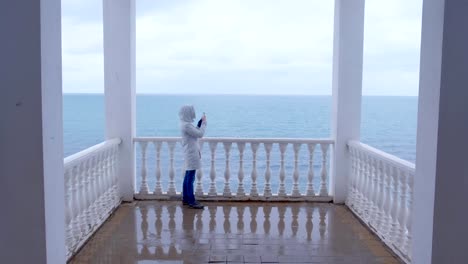 Frau-in-der-weißen-Jacke-schießt-Meerwellen-Video-auf-Smartphone-auf-schöne-Terrasse-mit-Meerblick.-Rückansicht.