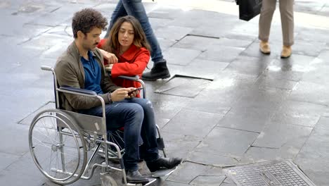 Mujer-arrodillada-cerca-de-su-novio-en-silla-de-ruedas-mirando-en-smartphone-al-aire-libre