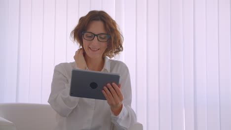 Closeup-portrait-of-adult-redhead-attractive-businesswoman-in-glasses-using-the-tablet-sitting-in-the-armchair-in-the-white-office