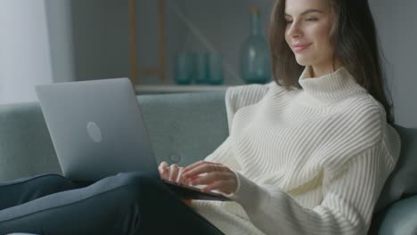 Schöne-junge-Frau-arbeitet-auf-Laptop-Computer,-während-auf-dem-Stuhl-sitzen.-Sinnliche-Mädchen-tragen-Pullover-funktioniert-auf-Notebook;-Studien,-Surfs-Internet,-Nutzt-Social-Media-beim-Entspannen-in-Cozy-Apartment