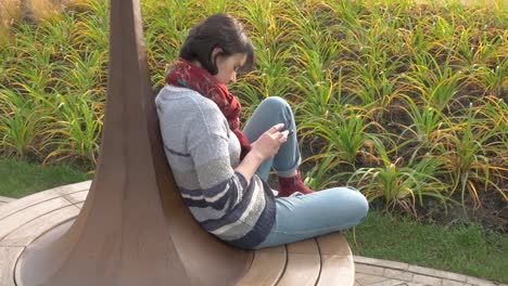 Caucasian-woman-sits-on-a-round-bench,-clicks-with-a-phone-in-her-hands