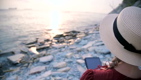 Close-up-View-from-the-back-An-attractive-young-girl-in-a-summer-red-dress-and-a-straw-hat-sits-on-a-stone-by-the-sea-at-sunset-and-watches-something-on-a-tablet.-Swipe-across-the-screen