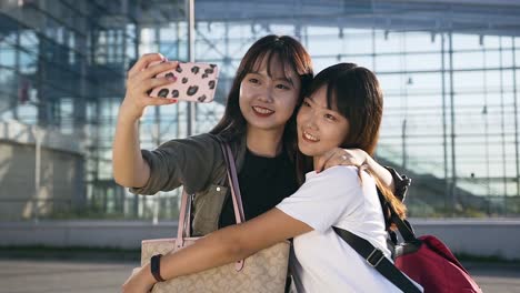 Smiling-elated-young-asian-women-in-modern-clothes-hugging-and-making-selfie-near-the-airport