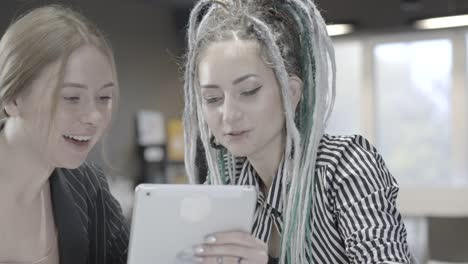 Two-positive-women-using-tablet-laughing-and-giving-high-five.-Close-up-of-positive-Caucasian-millennial-coworkers-celebrating-startup-success-in-office.-Teamwork-and-joy.