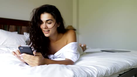 Junge-Frau-mit-Zelle-Smartphone-glücklich-lächelnde-schöne-Mädchen-Schlafzimmer-morgens-am-Bett-liegen