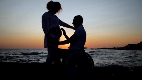 disabled-man-in-wheelchair-listens-to-abdomen-of-his-wife-in-evening,-love,-pregnant-female-with-disabled-husband