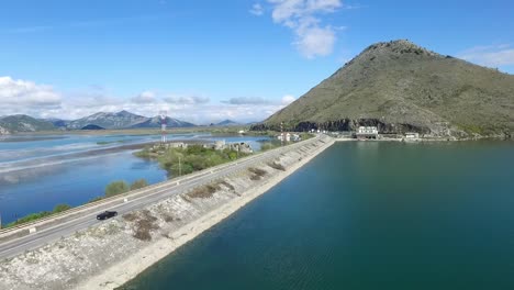 Black-car-driving-on-bridge-blue-sea,-mountains-and-sky-background.-Footage.-Car-going-on-a-road-near-the-river,-aerial-view.-Footage.-Aerial-view-at-small-forest-lake-and-line-of-highway-passing-near