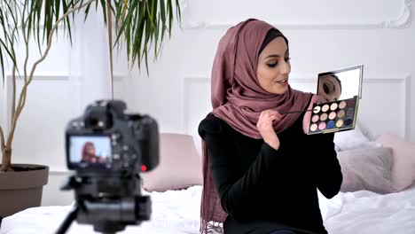 Young-beautiful-indian-girl-in-hijab-blogger-talking-on-camera,-smiling,-showing-a-new-purchase,-cosmetics,-Eyeshadow-Pallet,-home-comfort-in-the-background.-60-fps