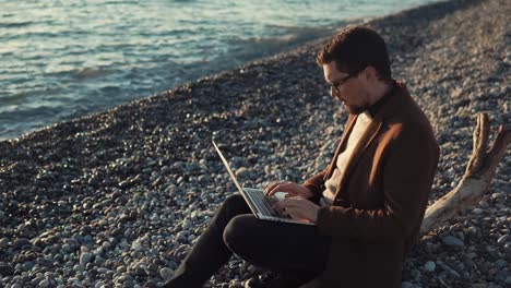 Bearded-man-is-typing-text-on-a-laptop,-sitting-on-a-seashore,-side-view