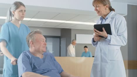 In-der-Klinik-Lobby,-Krankenschwester-schiebt-älteren-Patienten-im-Rollstuhl,-zwar-Arzt-spricht-mit-ihnen-mittels-Tablet-Computer.-Sauberes,-neues-Krankenhaus-mit-medizinischen-Fachpersonal.-Slow-Motion.