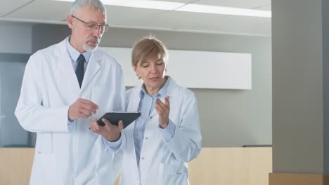 In-the-Hospital-Doctors-have-Discussion-while-Using-Tablet-Computer.-They're-Walking-through-the-Hallway.-In-the-Background-Patients-and-Medical-Personnel.-New-Modern-Fully-Functional-Medical-Facility.