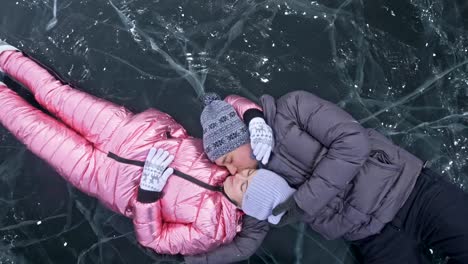 Young-couple-has-fun-during-winter-walk-against-background-of-ice-of-frozen-lake.-Lovers-lie-on-clear-ice-with-cracks,-have-fun,-kiss-and-hug.-View-from-above.-Honeymoon.-Love-story.