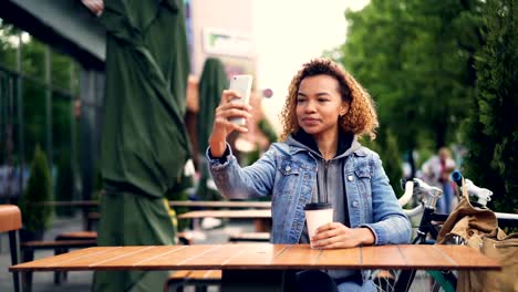 Unvorsichtige-afroamerikanische-Frau-froh-Tourist-nimmt-Selfie-mit-Smartphone-im-Straßencafé-mit-Take-Away-Kaffee-am-Tisch-sitzen,-Fahrrad-und-Rucksack-sind-sichtbar.