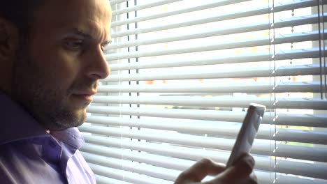 Young-businessman-standing-with-smartphone-by-window-in-the-office.-Close-up,-serious-man-looks-out-the-window-and-uses-a-smartphone.-Thoughtful,angry,-problem,-social-network.