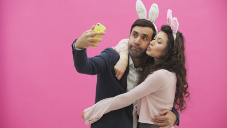 Young-tender-pair-standing-on-a-pink-background.-Looking-at-the-phone-while-doing-selfie-on-the-mobile-phone-with-the-ears-of-a-pink-rabbit-on-the-head.-Happy-family-is-preparing-for-Easter