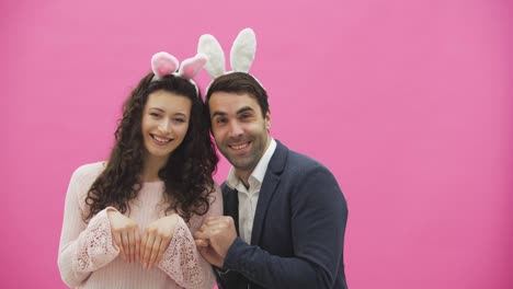 Beautiful-young-couple-standing-on-pink-background.-During-this-time,-they-recreate-the-movements-of-hares,-placing-their-hands-like-a-rabbit.-Together-with-pink-barking-ears-on-the-head.-Happy-family-is-preparing-for-Easter.