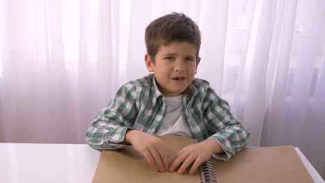 Visually-impaired-small-boy-reading-braille-book-with-symbols-font-for-blind-sitting-at-table