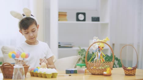 Creative-boy-is-colorizing-an-Easter-egg-with-a-help-of-paint-brush.,-wearing-bunny-ears,-then-he-takes-a-wooden-basket-and-shows-how-many-coloured-Easter-eggs-there-are.