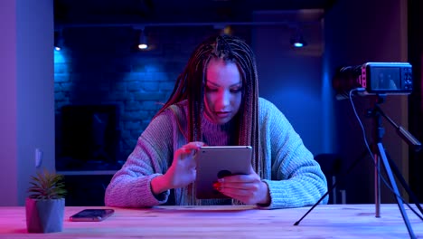 Closeup-shoot-of-young-attractive-female-blogger-with-dreadlocks-texting-on-the-tablet-smiling-cheerfully-streaming-live-with-the-neon-background-indoors