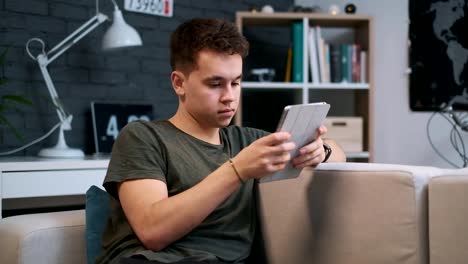 Portrait-view-of-a-young-guy-sitting-on-the-couch-and-sufring-the-web-on-his-tablet