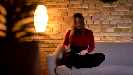 Closeup-shoot-of-young-pretty-caucasian-female-texting-on-the-tablet-then-looking-at-camera-with-thoughtfulness-sitting-on-the-couch-indoors