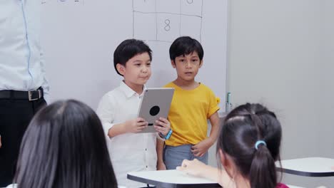 Niño-explicando-el-tema-al-estudiante-en-el-salón-de-clases.-Niño-usando-tableta-para-ayudar-en-su-tema.-Concepto-de-Educación-y-Tecnología.