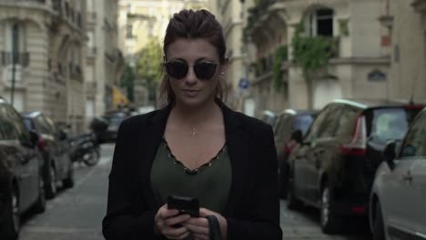 Attractive-caucasian-classy-woman-with-sunglasses,-freckles,-piercings,-black-jacket-and-red-hair-writing-a-text-message-smiling-walking-through-the-street,-during-sunny-day-in-Paris.-Slow-motion.