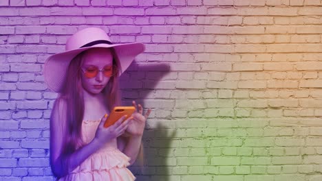 Teenager-girl-browsing-mobile-phone-on-brick-wall-background-in-colorful-light.-Young-girl-using-smartphone-for-surfing-in-social-networks-on-multicolored-brick-wall-background.