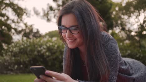 Portrait-of-an-attractive-smiling-young-woman-lying-in-park-texting-messages-on-smartphone