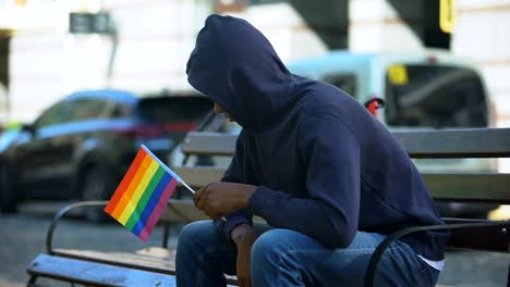 Black-man-in-hood-sitting-on-bench-with-lgbt-minority-flag,-preconceptions